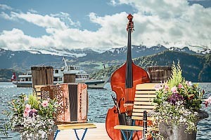 Foto mit Bassgeige, Akkordeon und Klarinette vor einem stimmungsvollem Panorama mit See, Bergen und Schiff.